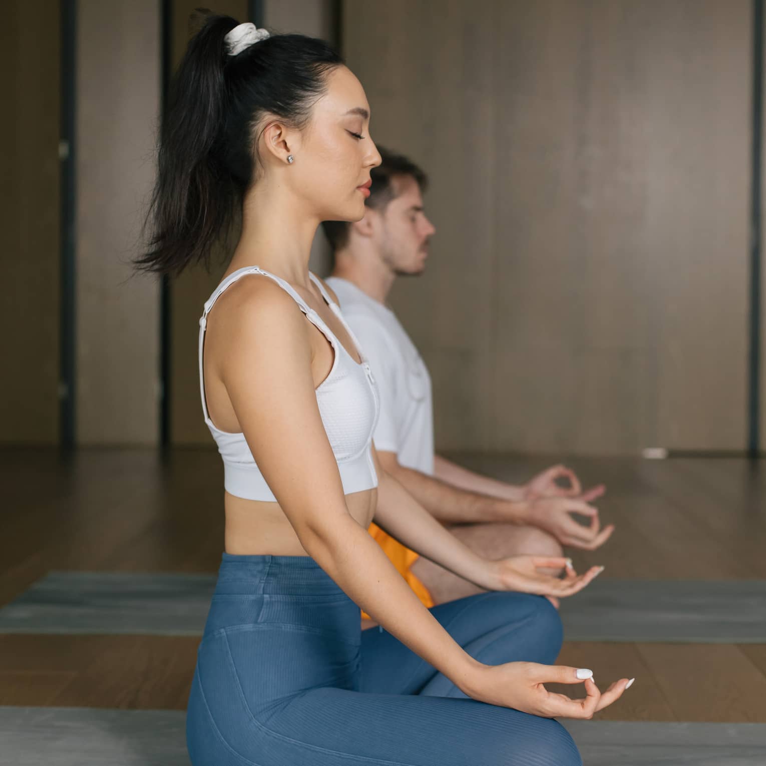 Woman with dark hair tied up in a ponytail and wearing a white tank top and blue yoga pants sits cross-legged on a yoga mat with her hands on her knees and eyes closed, a man wearing a white t-shirt and orange shorts does the same behind her