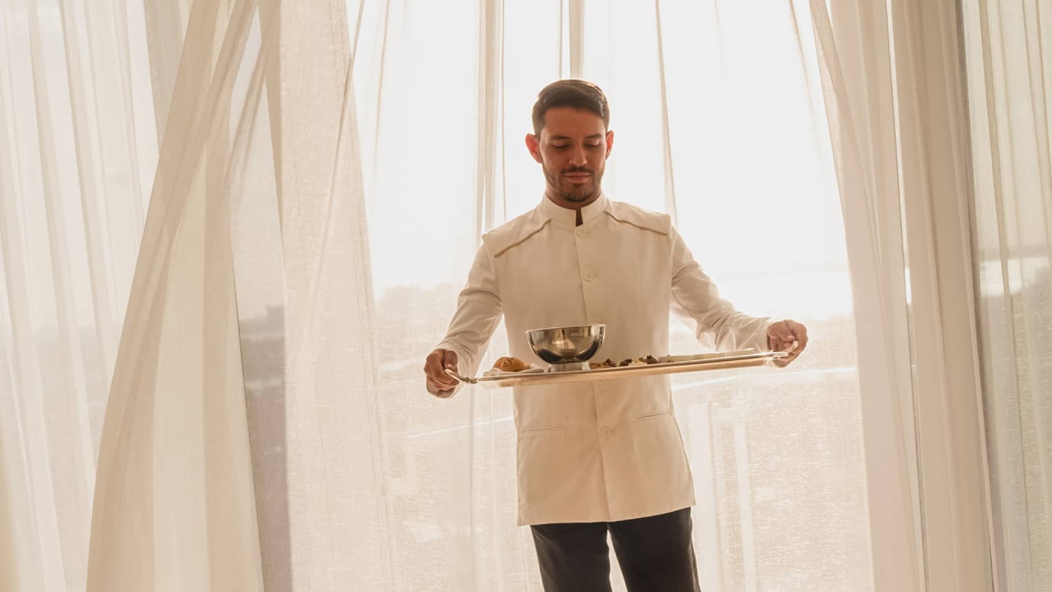 Hotel staff member carrying a tray with food for in-room dining, standing in front of sheer curtains with sunlight streaming through