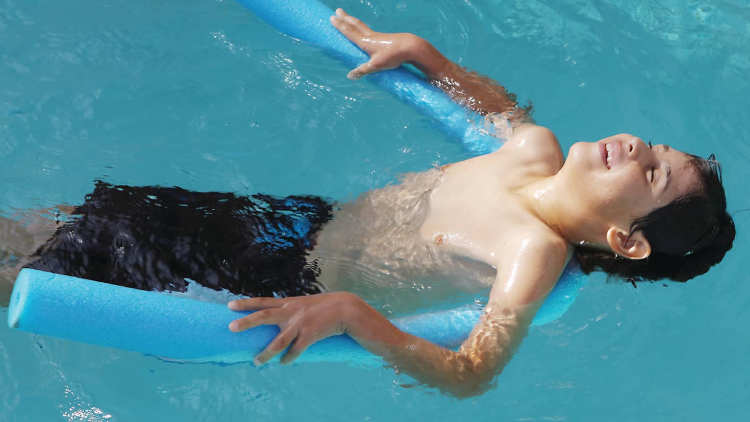 Boy leans back, floats on blue foam pool noodle in outdoor swimming pool