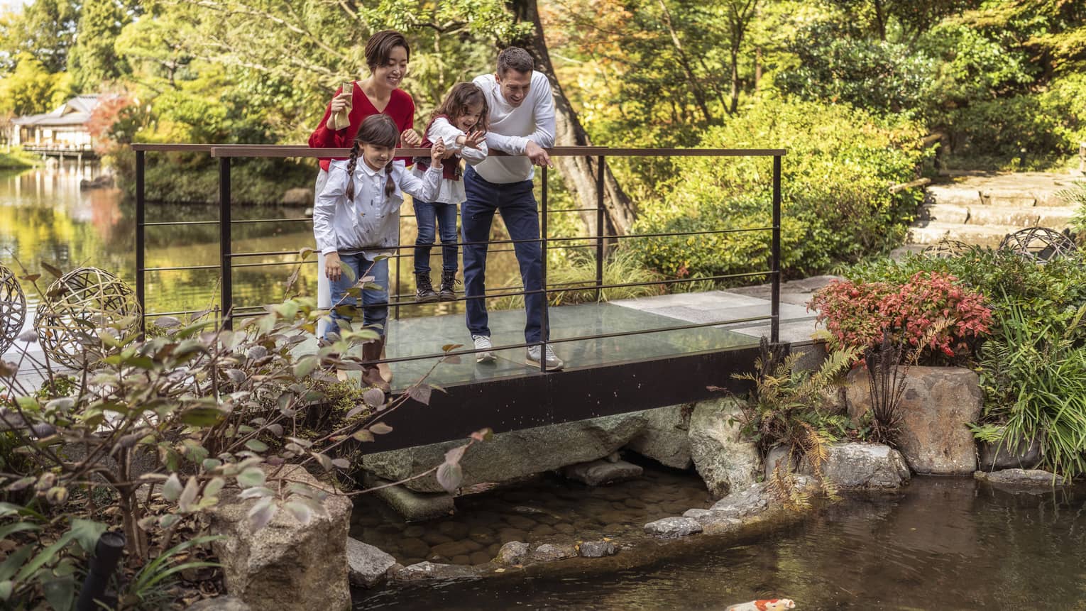 Parents and young son and daughter feed koi fish on foot bridge over pond