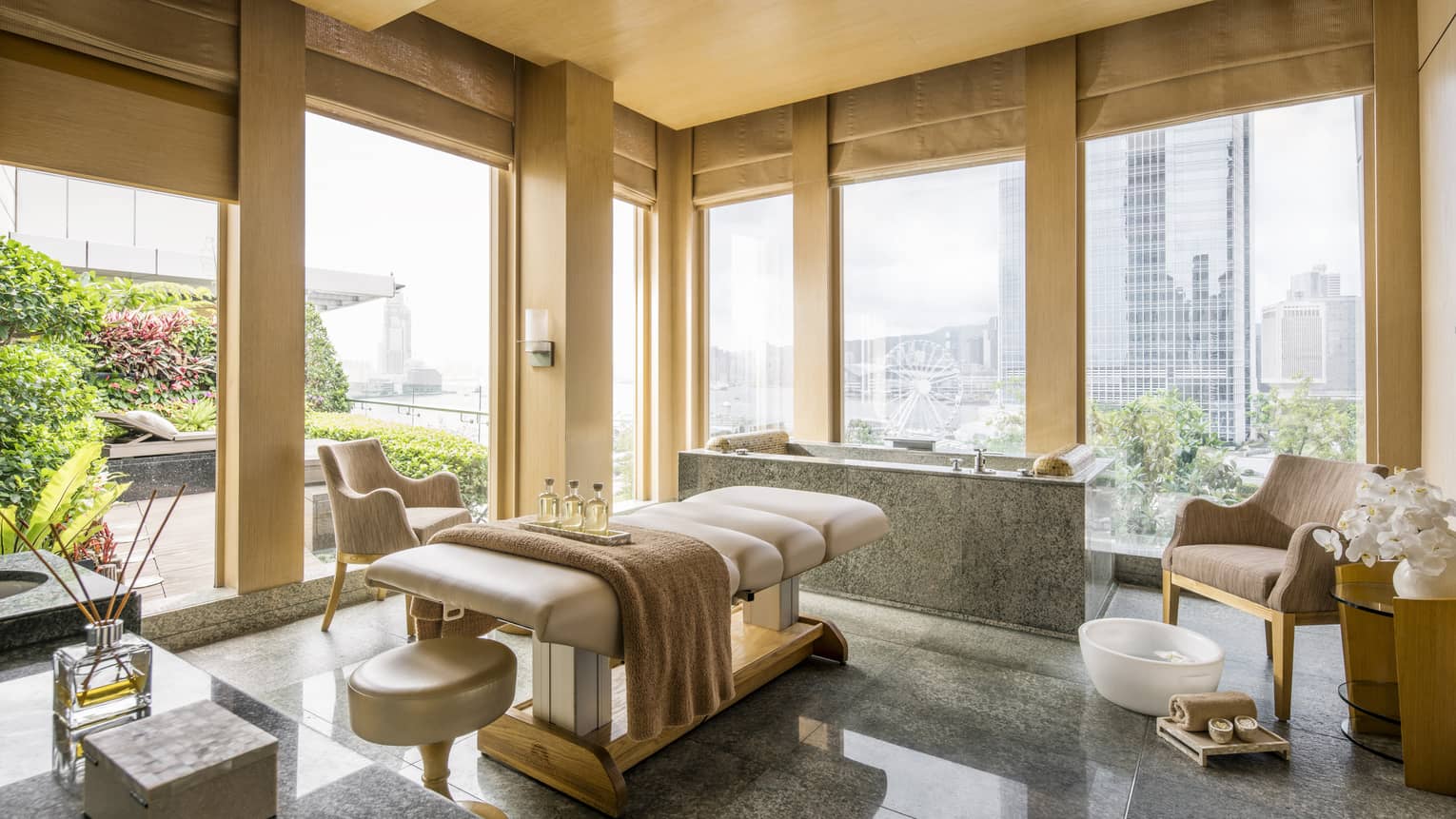 A serene spa room with a massage table, towels, and a city skyline view. Comfortable chairs and a foot bath are nearby.