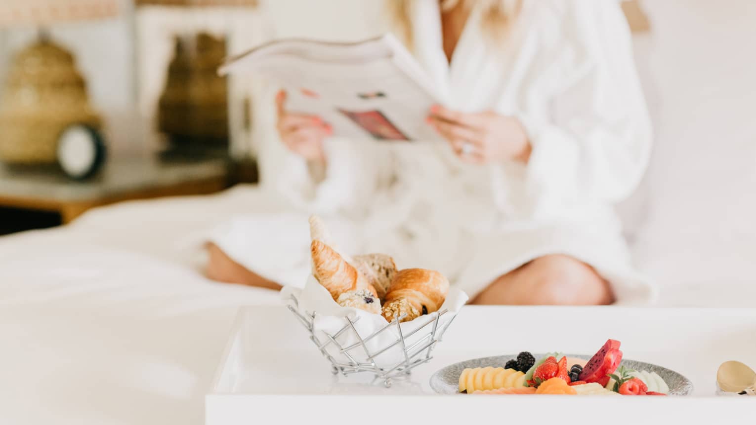 Pastries, fruit on in room dining tray, woman wearing white bath robe reads in background
