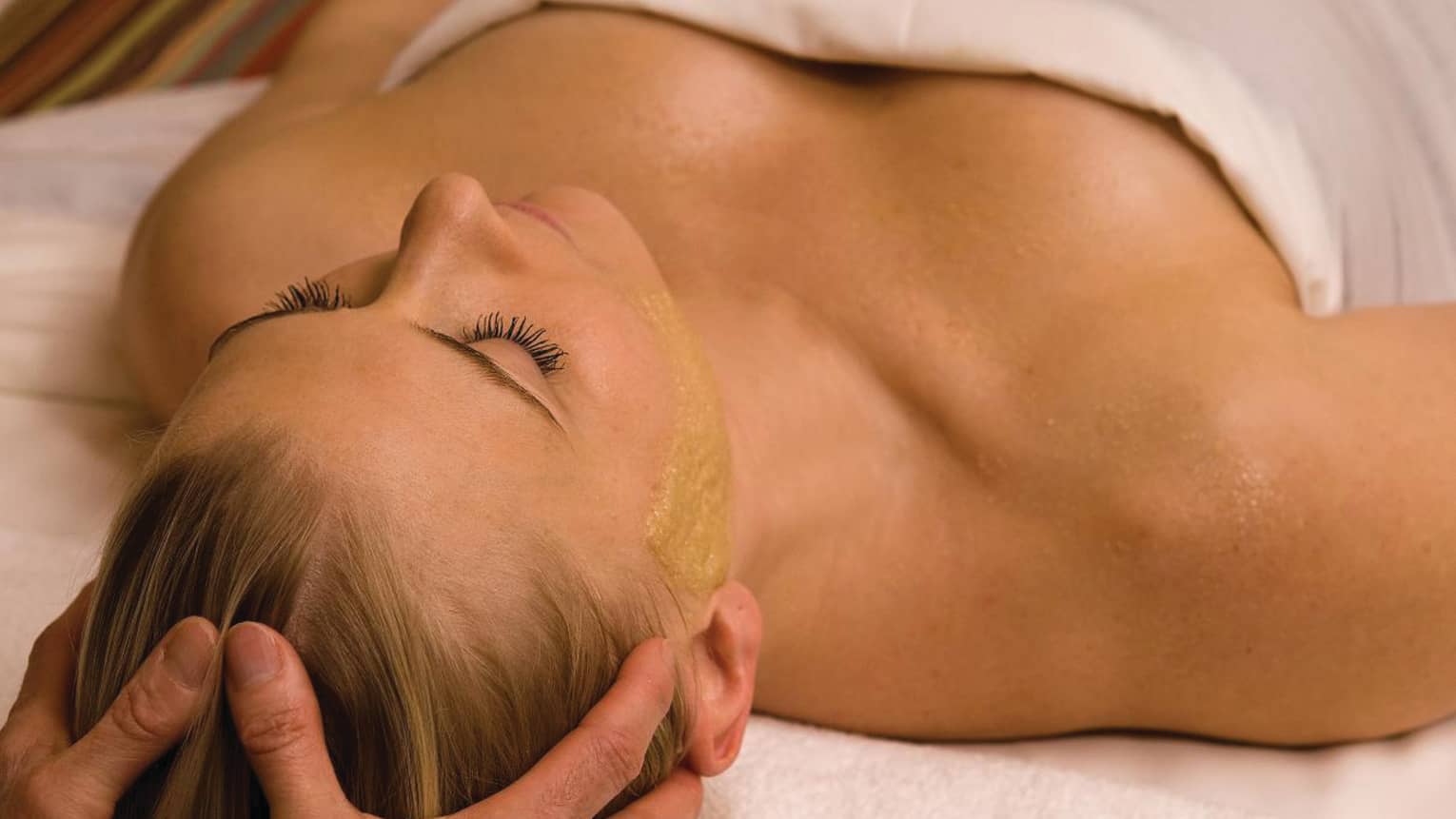 Woman wrapped in white sheet lies on spa table as hands massage her hair