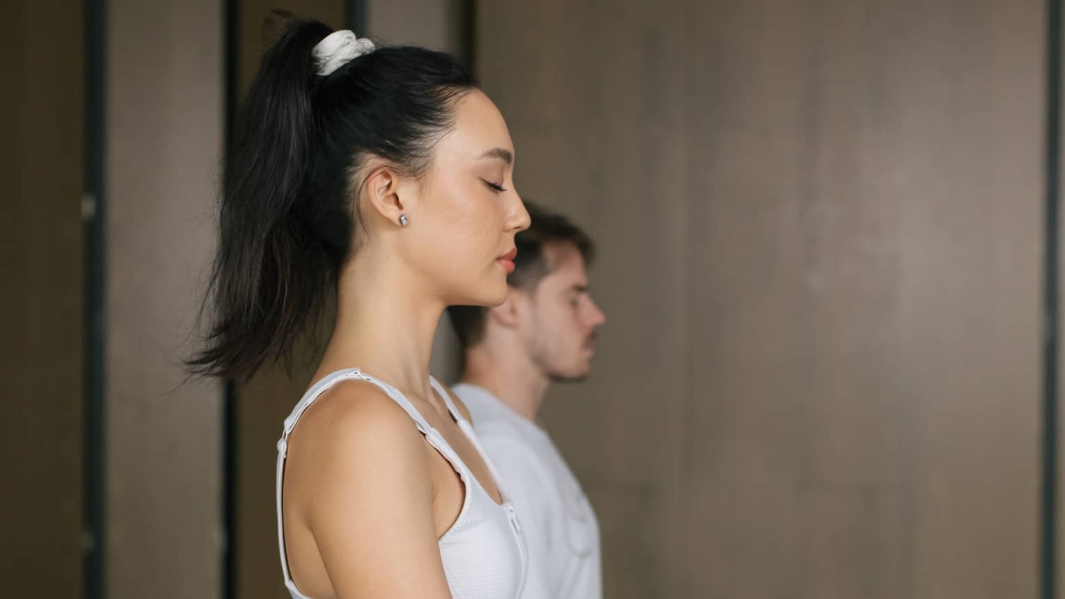 Woman with dark hair tied up in a ponytail and wearing a white tank top and blue yoga pants sits cross-legged on a yoga mat with her hands on her knees and eyes closed, a man wearing a white t-shirt and orange shorts does the same behind her