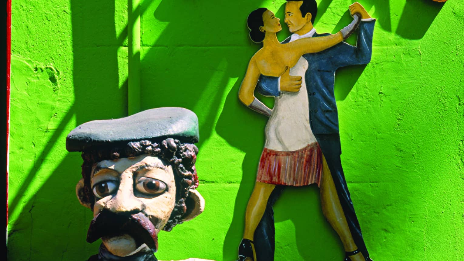 The top part of a marionette folk art statue; behind, a bas relief on a neon-green wall of a dancing couple in tight embrace.