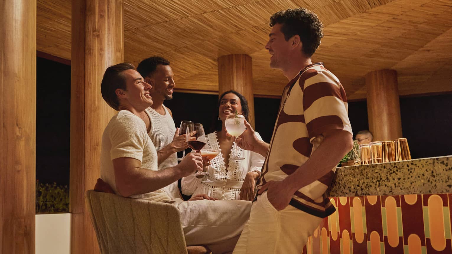 Four guests in stylish summer clothes hold cocktails and socialize next to a terazzo-topped bar in a round wooden pavilion.