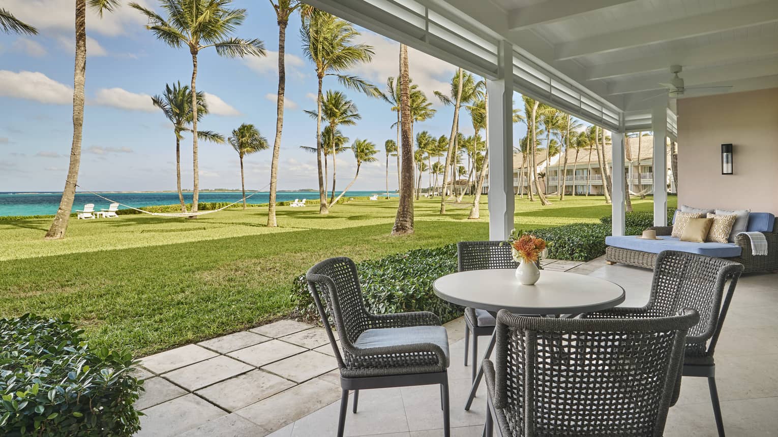Outdoor patio with a round dining table and chairs overlooking a lawn, palm trees and ocean 