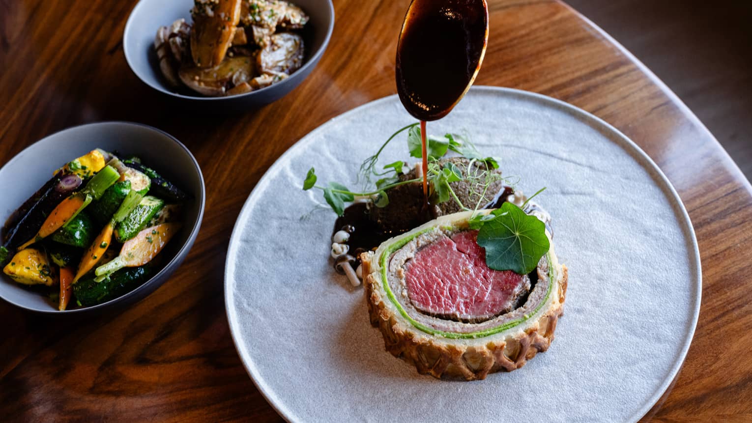 Slice of beef Wellington plated on a white dish with greens on the side plus a bowl of sliced vegetables and a bowl of mushrooms