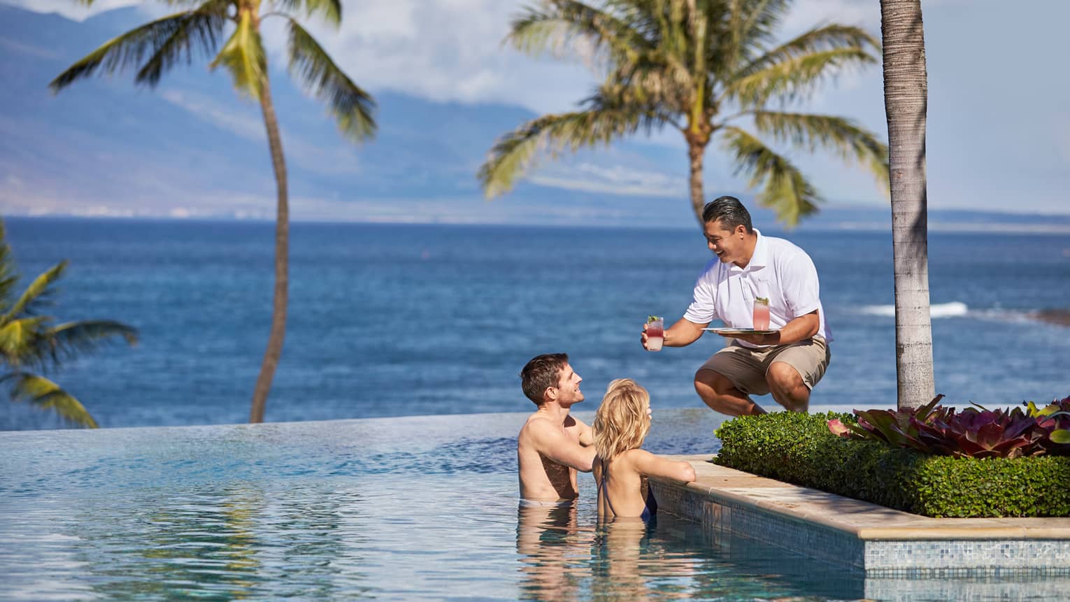 Server in white uniform with tray passes a cocktail to man and woman at edge of infinity swimming pool