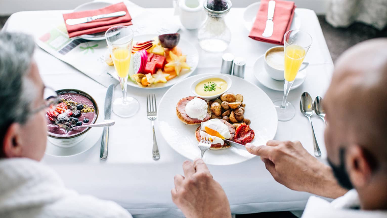 Two men eating room service breakfast while looking out a window, it includes eggs, sausage, acai bowl and orange juice.