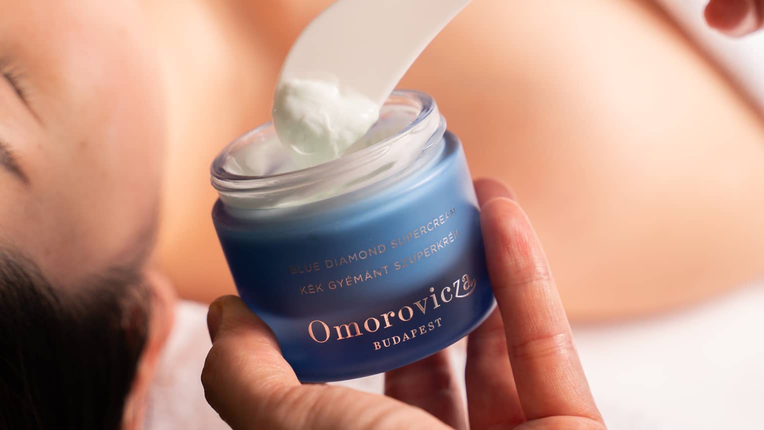 Hand dips spatula into jar of Omorovicza face cream in preparation for woman’s facial