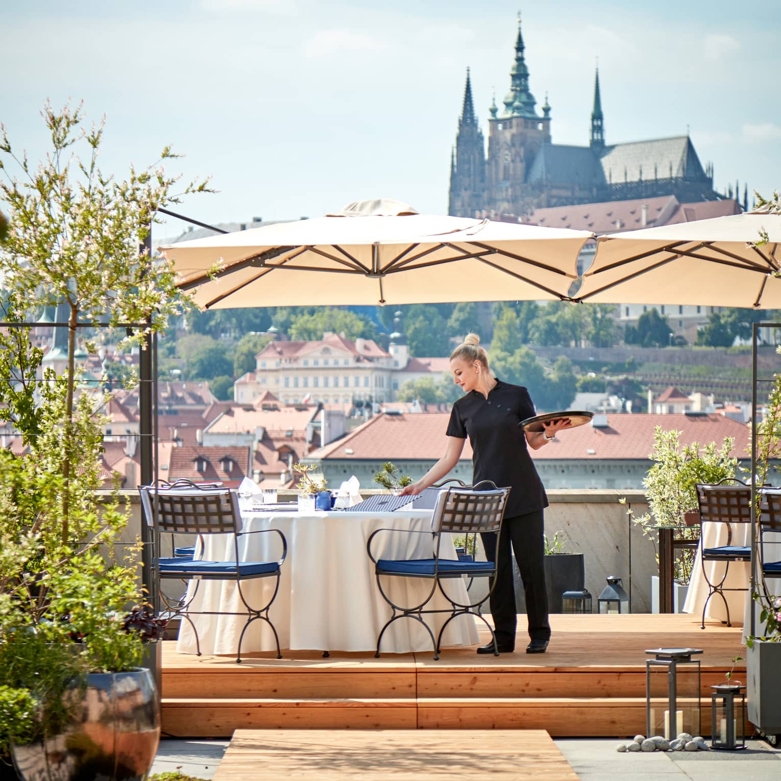 Server with tray in hand beside table with umbrella on rooftop restaurant, Prague Castle in view