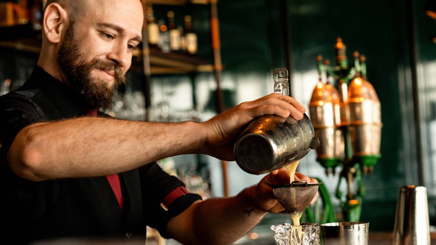 A smiling bartender pours a milky liquid through a strainer into a sculpted blown-glass tumbler, in a warm bar setting.