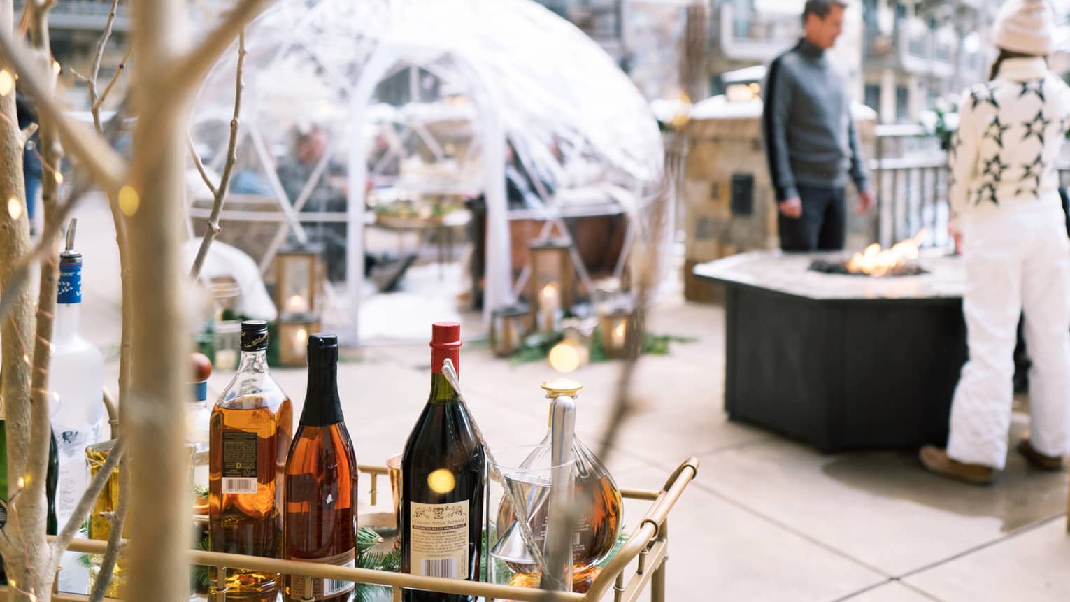 Three guests lounge on an outdoor patio with a fire pit, dining igloo and bar cart