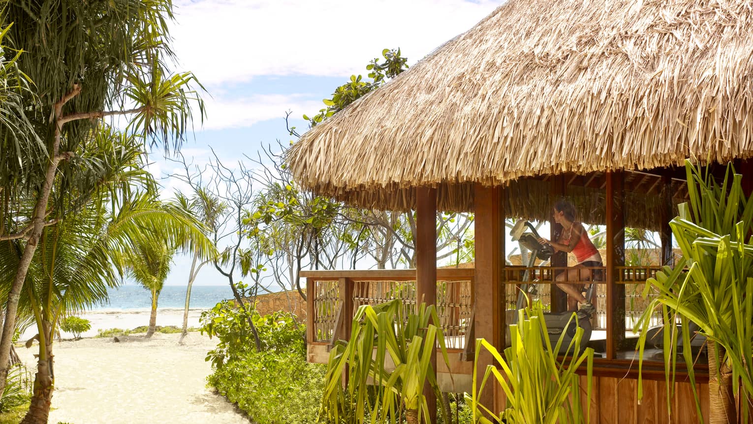 Woman on exercise bike in thatched-roof gym looks out at beach