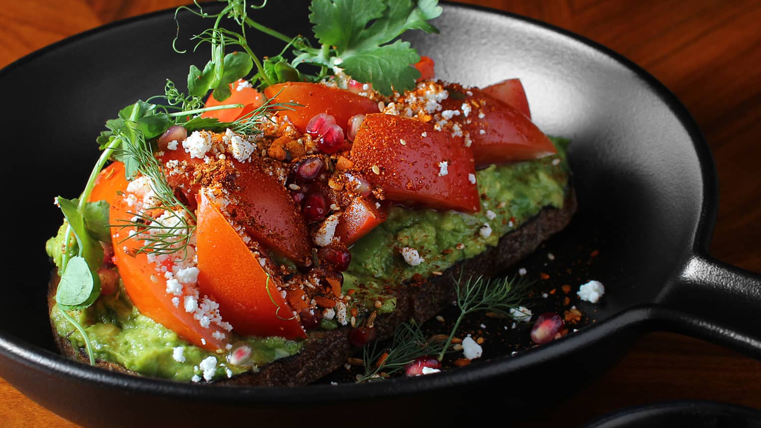 Avocado toast with tomatoes and herbs on a black plate.