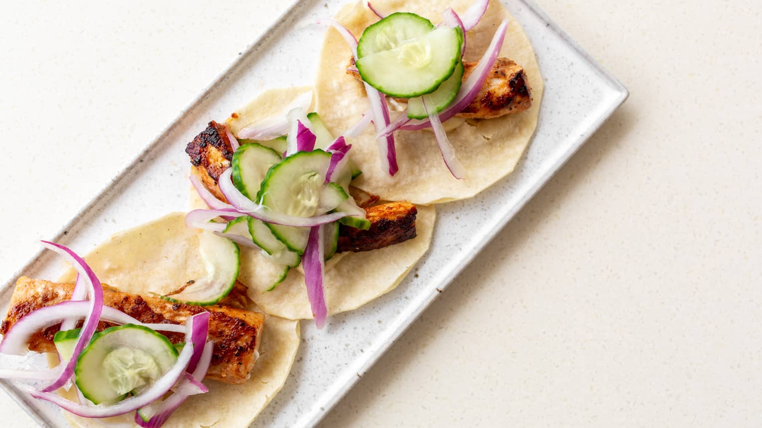 Three fish tacos lined up on a white rectangular dish, each topped with red onion slices and cucumber