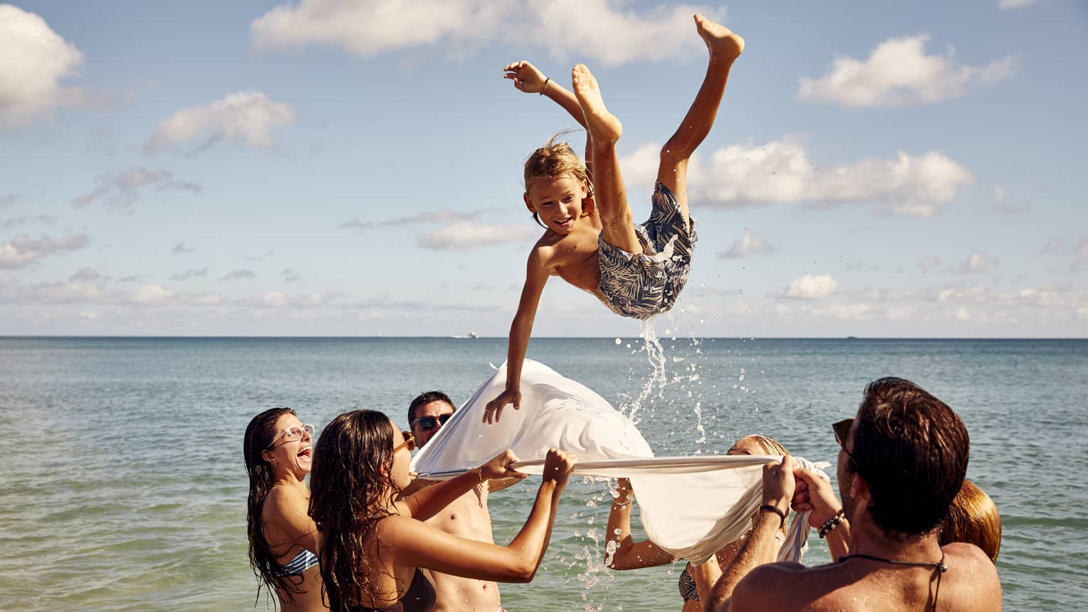 A group enjoys a sunny beach day, tossing a child into the air with a white sheet. The child smiles mid-air against clear skies.