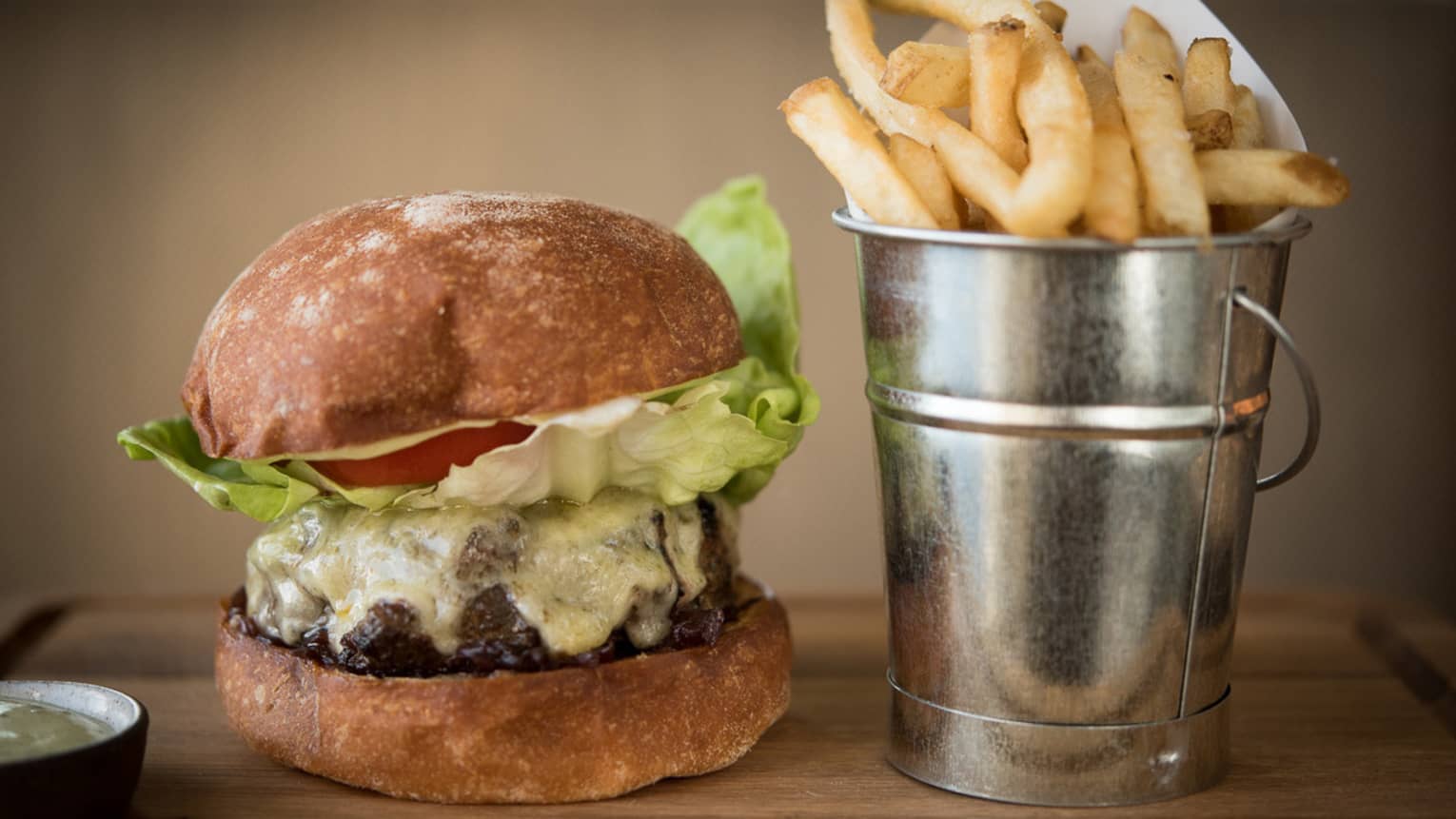 Thick beef burger with melted cheese on artisan bun beside silver tin with French fries