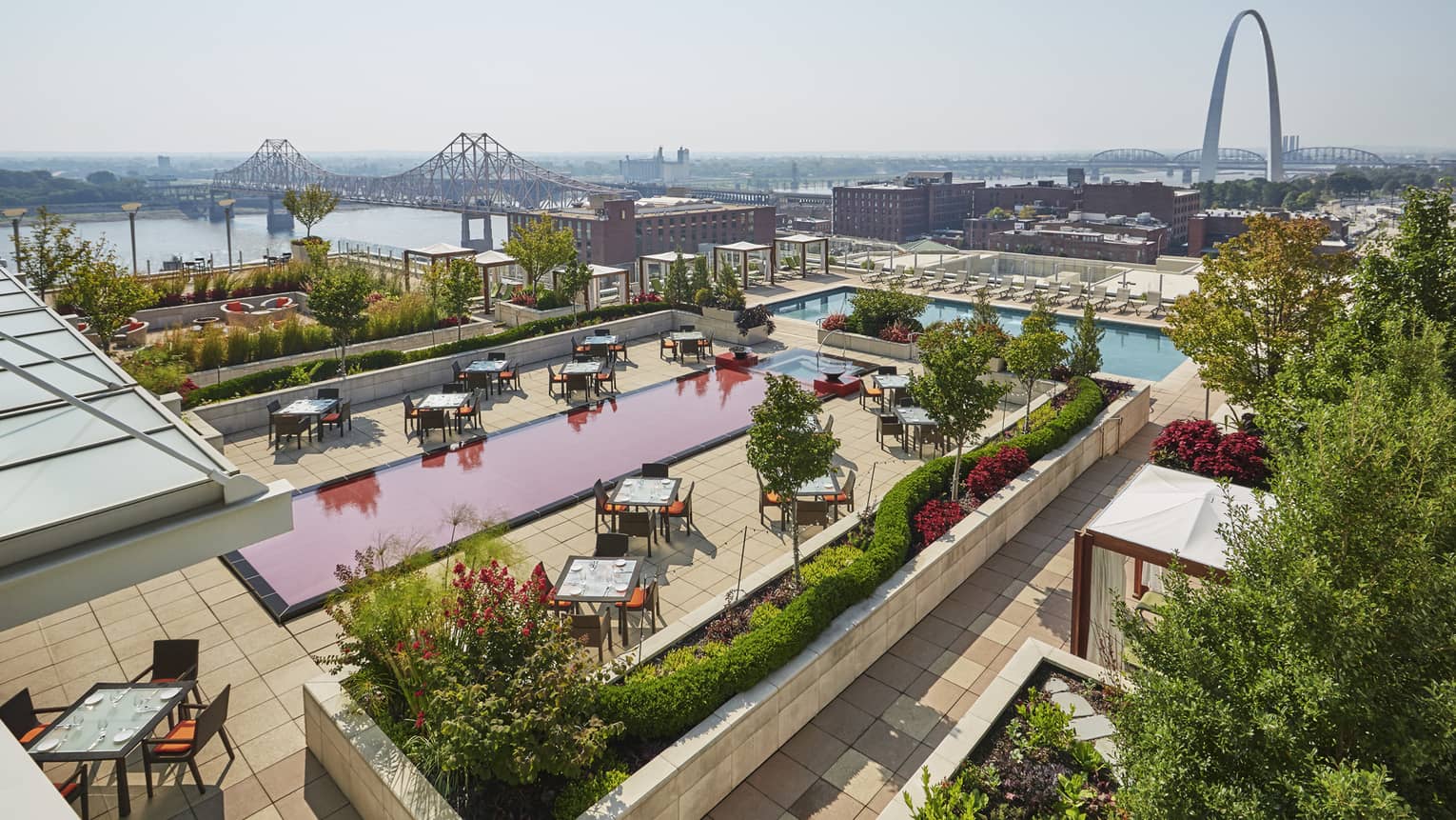 Hotel rooftop terrace with dining tables and a red and blue swimming pool, overlooking a cityscape, river and the Gateway Arch in the distance