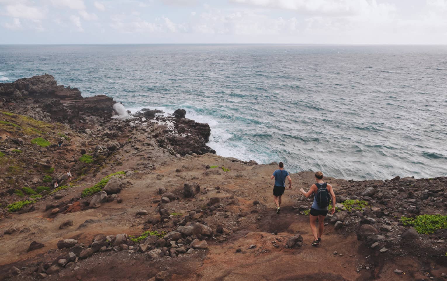Two people hike down dirt-covered path, rocks towards ocean