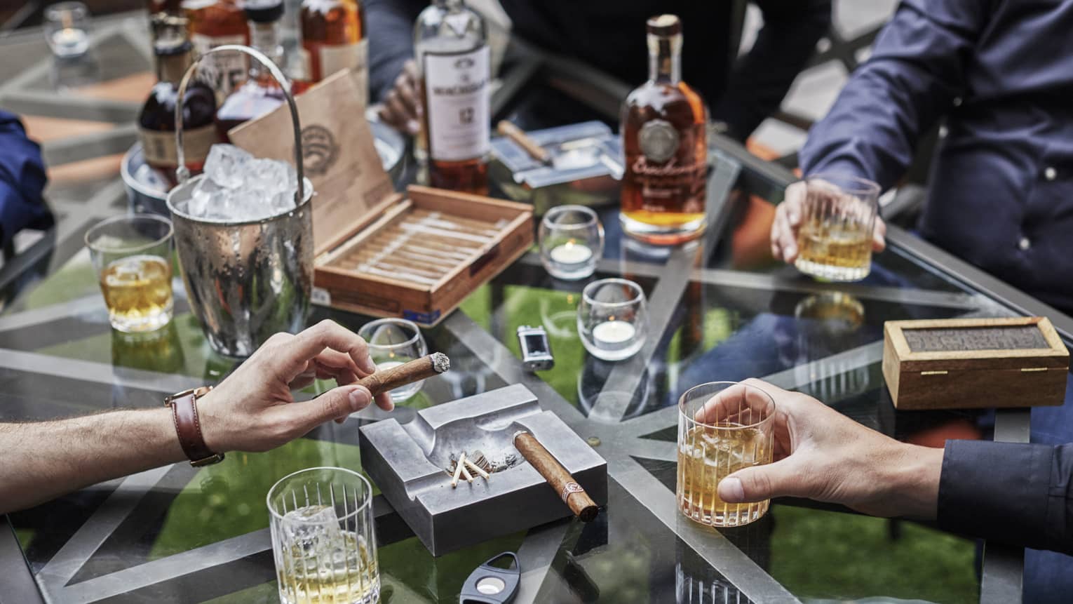 Bachelor party scene with men's hands holding rock glasses with liquor and thick cigars at glass table