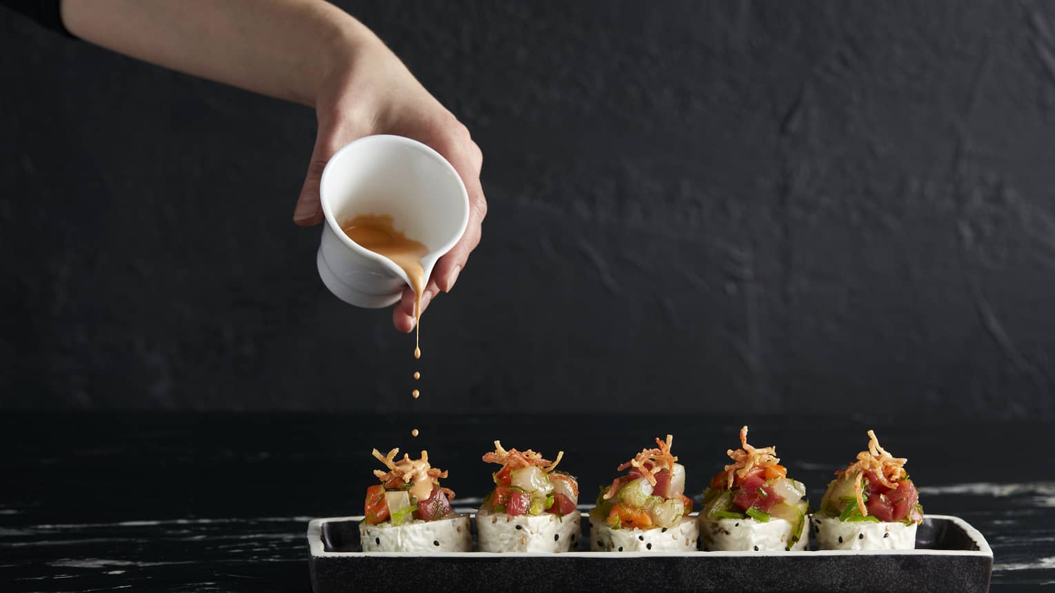 Five colourful sushi rolls set against a black background, hand pouring drops of orange-coloured sauce from a small carafe.