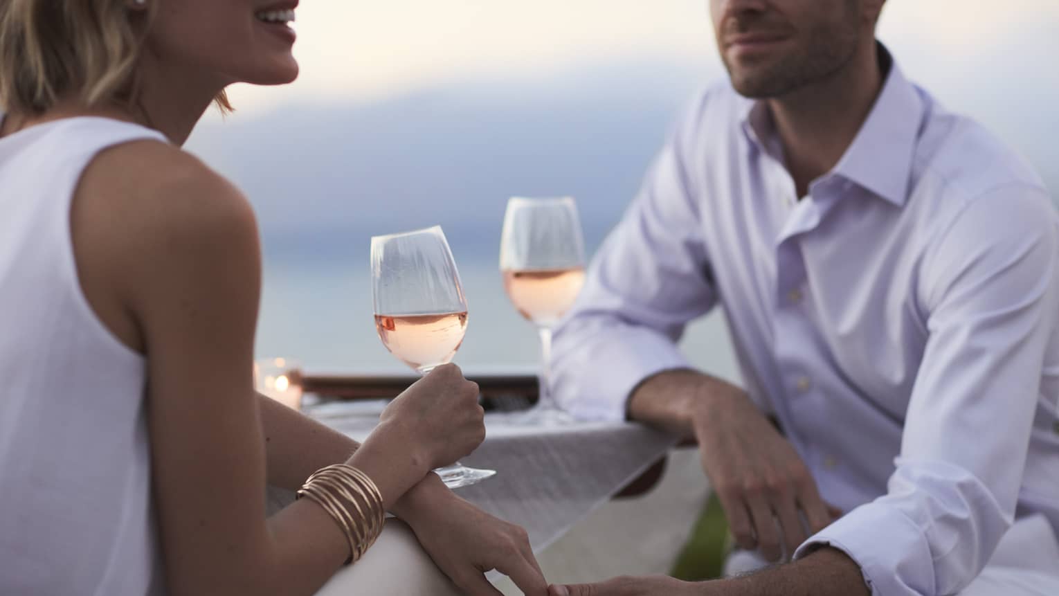 Close-up of woman and man in white, sitting and holding glasses of rose wine at dusk