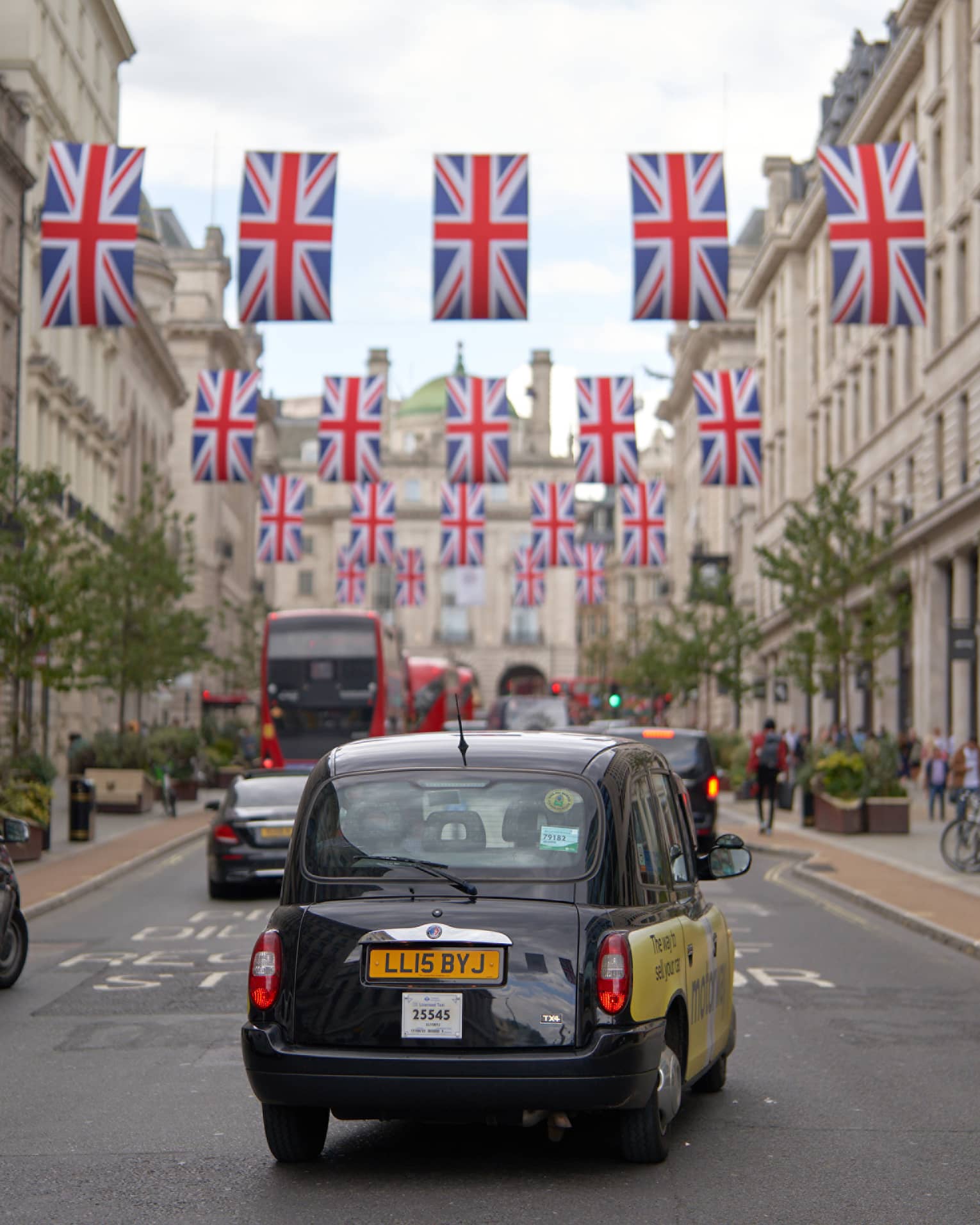 Rear view of London-style black cab, double-decker buses and other cars on road strung across with rows of Union Jack flags.