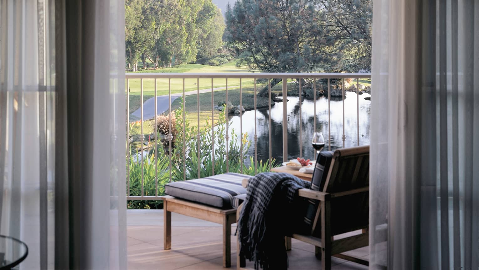 Patio chair with blanket on arm and wine glass on table, looking past railing to pond, garden, trees