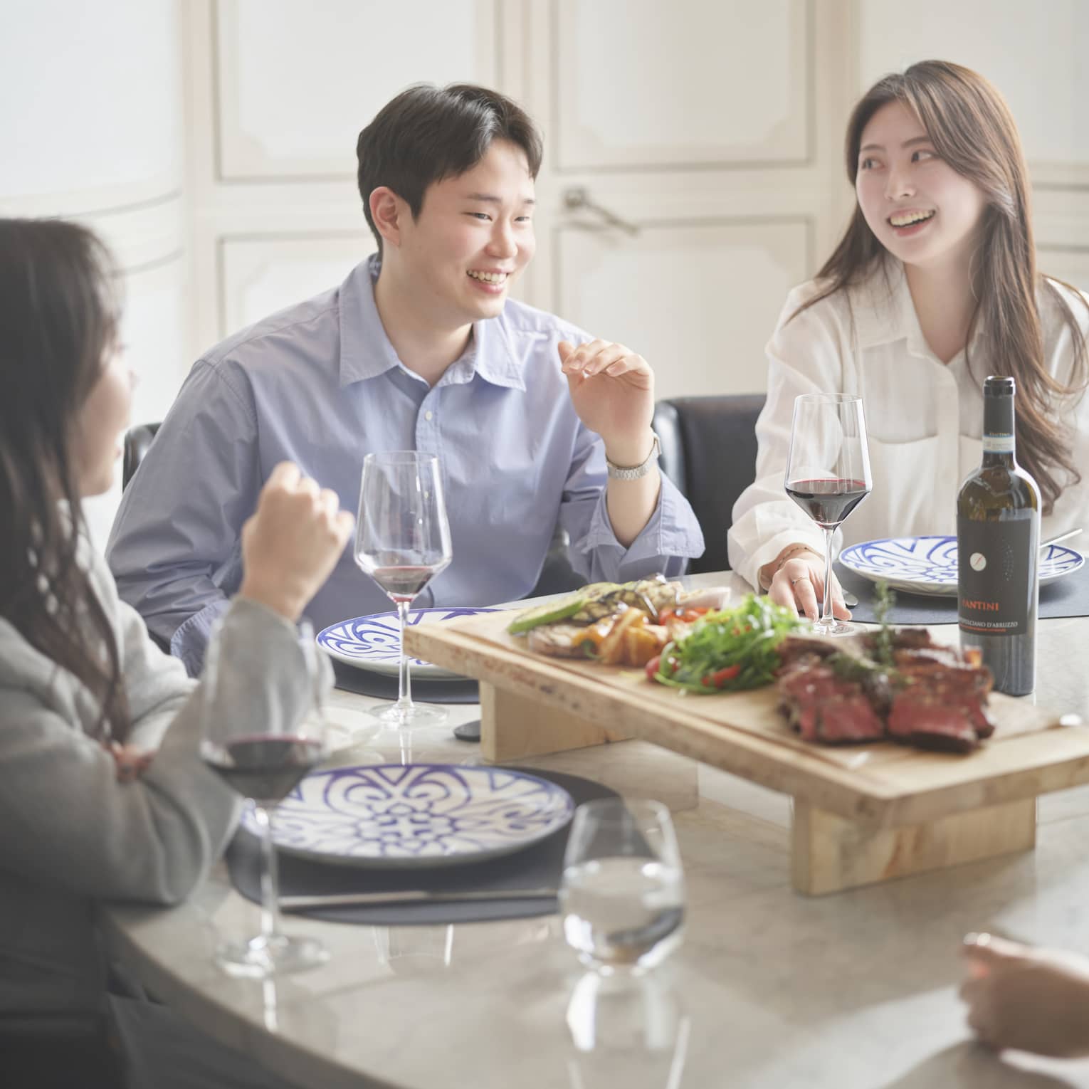 Group of friends sit around a table for a meal together, glasses of red wine and wooden display of meat and vegetables surrounding them