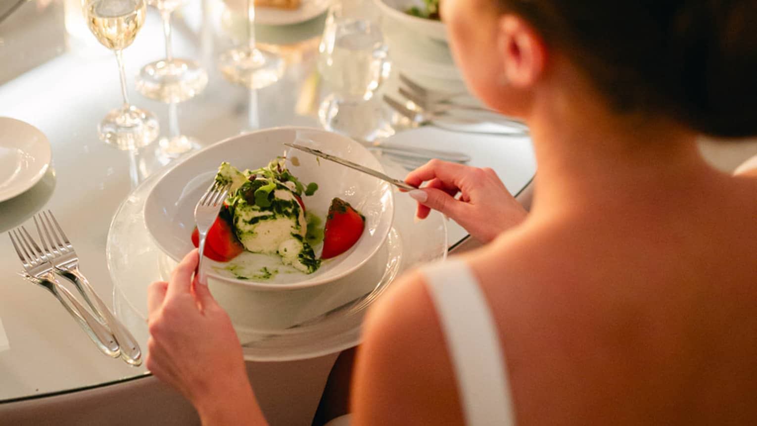 Person in bridal gown sits at round table set for dinner, fork and knife posed to dig into a meal