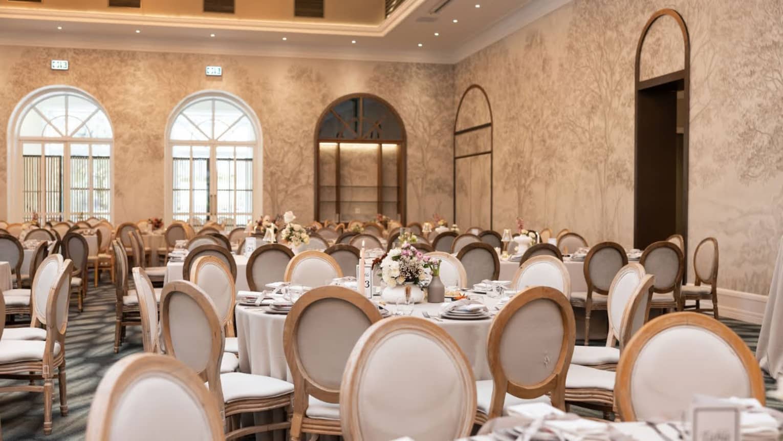 A spacious ballroom filled with round dining tables and chairs for an event. Large arched windows let in natural light, and ceiling lighting adds to a bright, warm ambience. Walls are warm and neutral in tone, with subtle nature-themed designs.