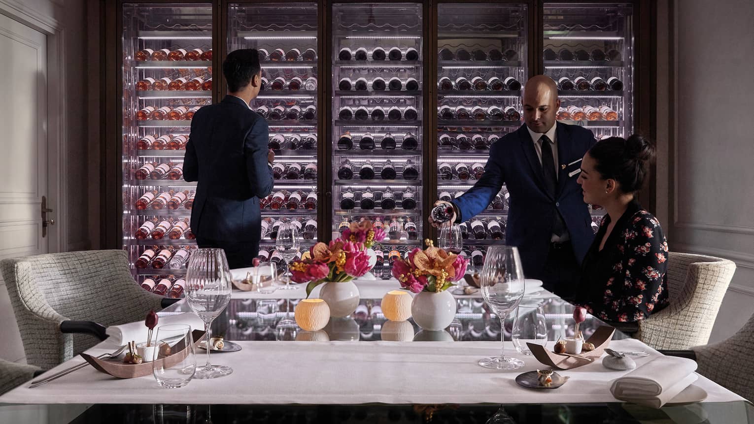 Man scans glass cellar as waitstaff pours wine in glass for woman at table
