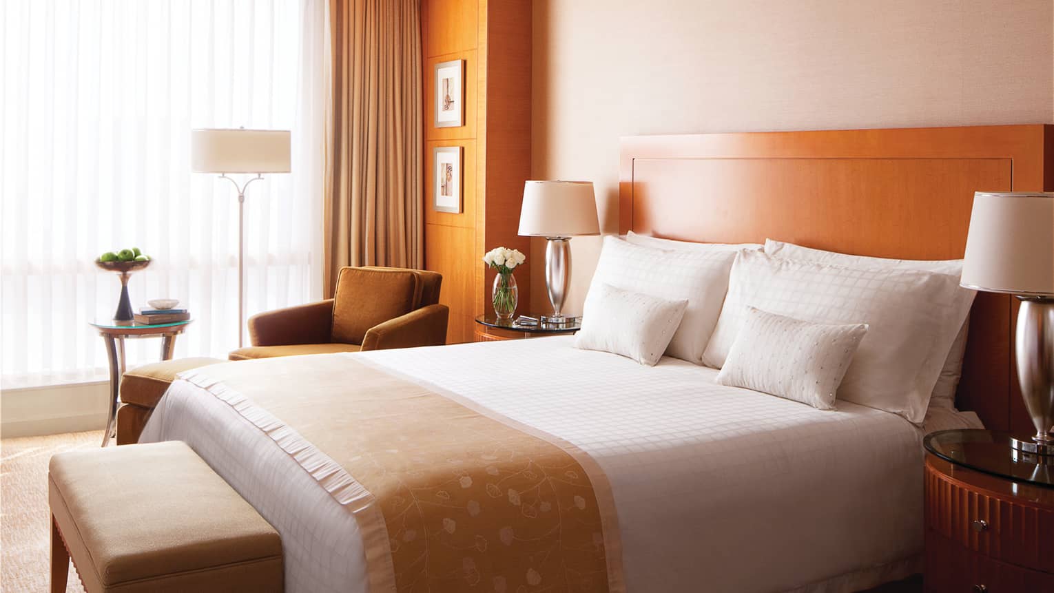 Deluxe Room hotel bed with embroidered gold sash, bench, armchair by window, sheer curtains