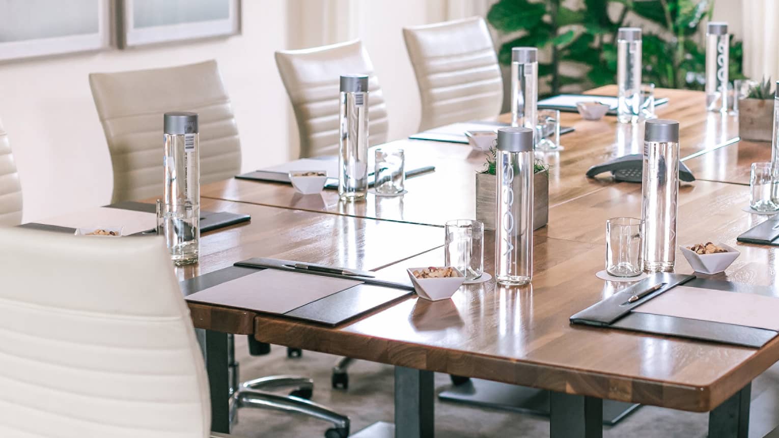 A rectangular boardroom table set with clear glass water bottles and black folders in a naturally-lit room