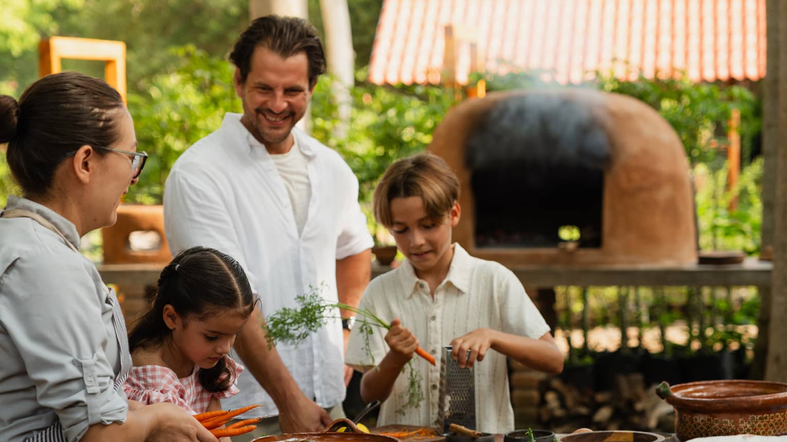 Adults and children standing around an outdoor dining table with various dishes and utensils on it, while an outdoor oven runs in the background.