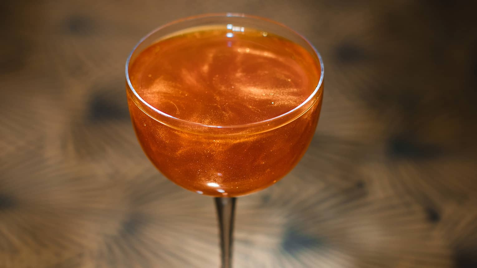 Bright-orange clear cocktail in wine glass atop bronze patterned table