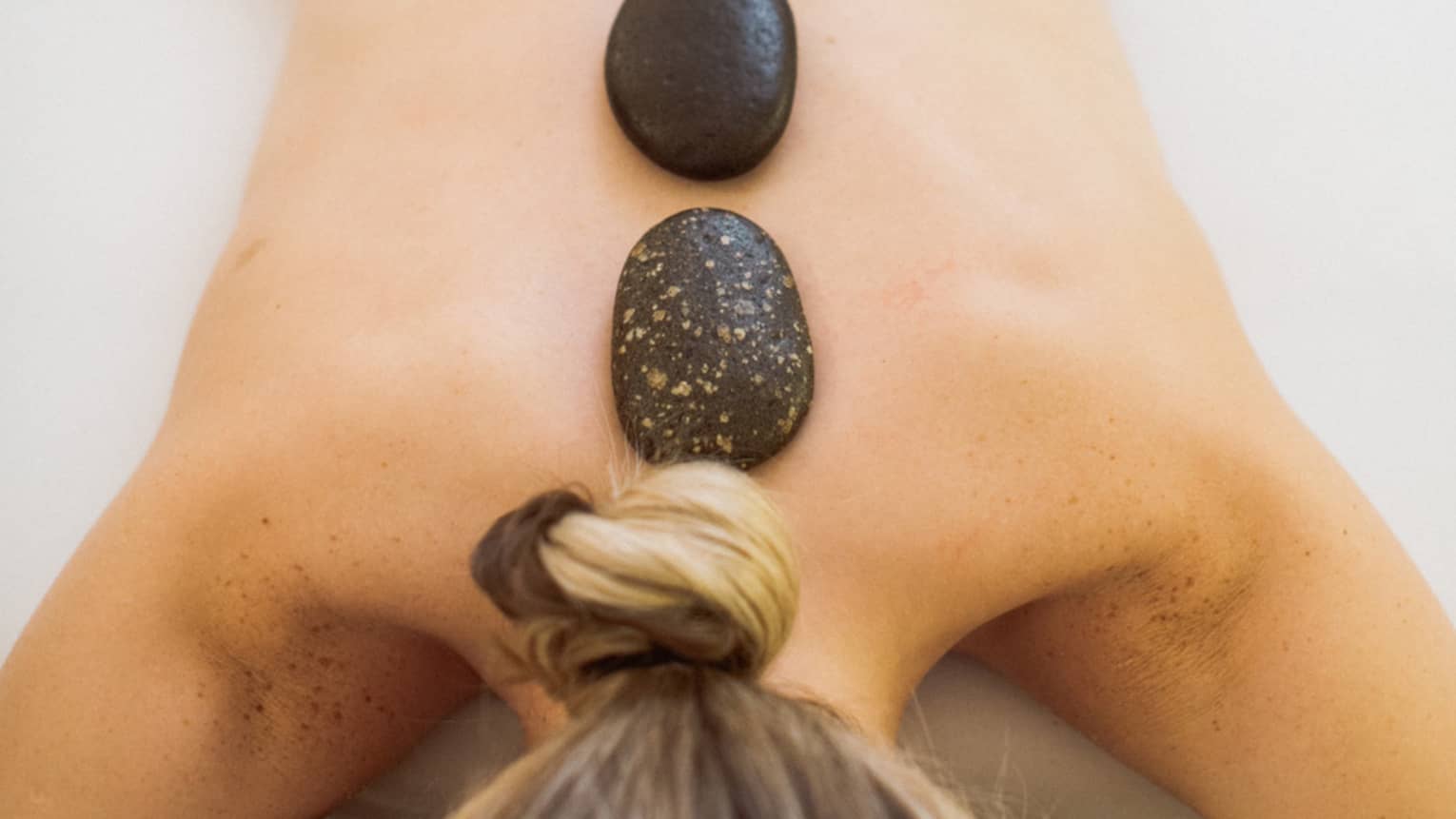 Woman lays under white towel on massage table, five black stones placed along her spine