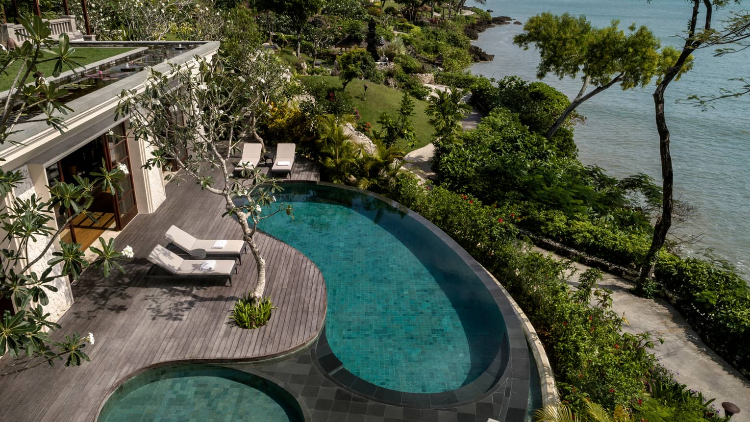 Looking down at Imperial Villa swimming pool and whirlpool on deck with white lounge chairs, garden path and ocean below