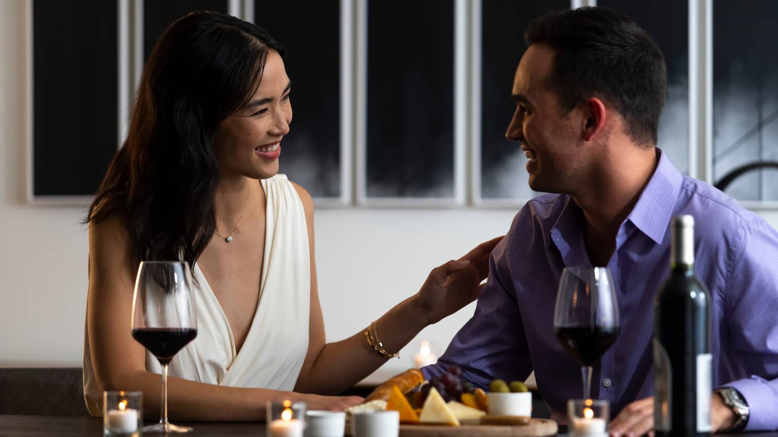 A couple exchanges smiles as they enjoy a cheese platter and a bottle of wine.