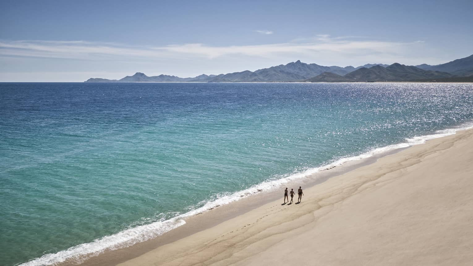 Three people walk on a white-sand beach with mountains in the distance