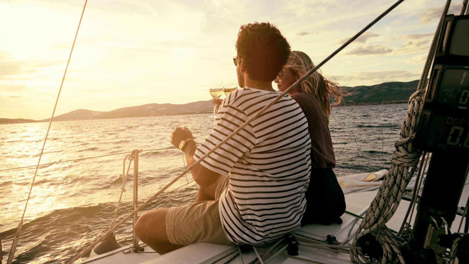 Man in striped T-shirt and woman with long hair toast with wine glasses on side of luxury yacht at sunset