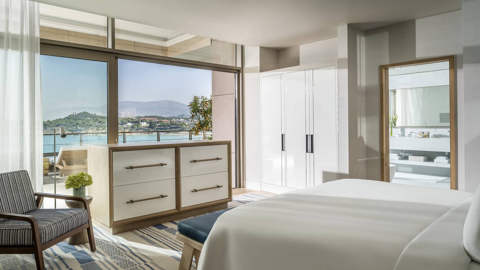 Arion Royal Suite with white King bed, white corner wardrobe, striped area rug, grey arm chair, doorway to bathroom, ocean views