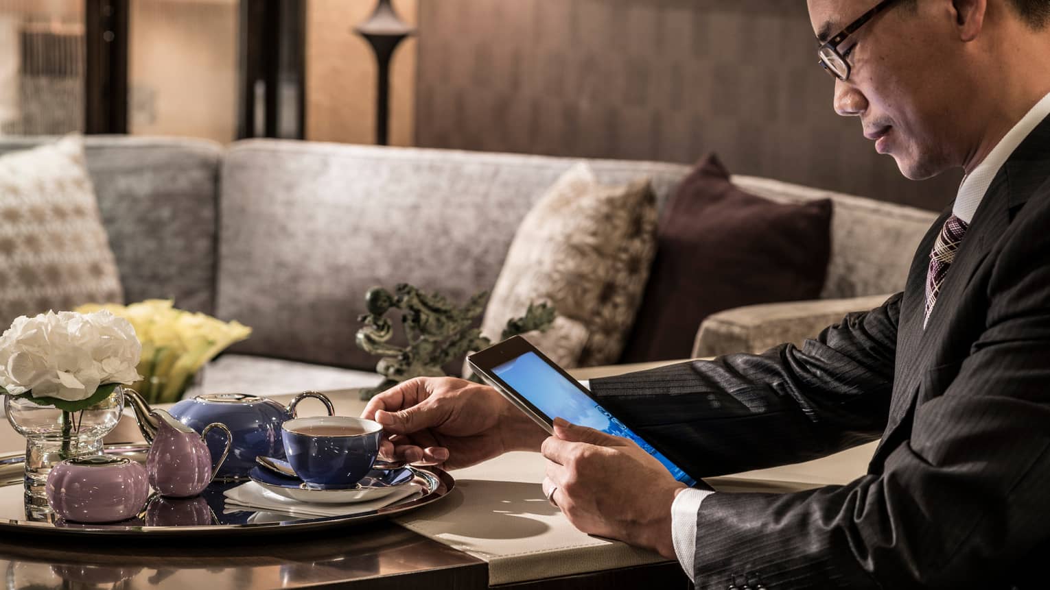 Man in suit holds tea cup on tray, reads iPad by elegant purple sofa