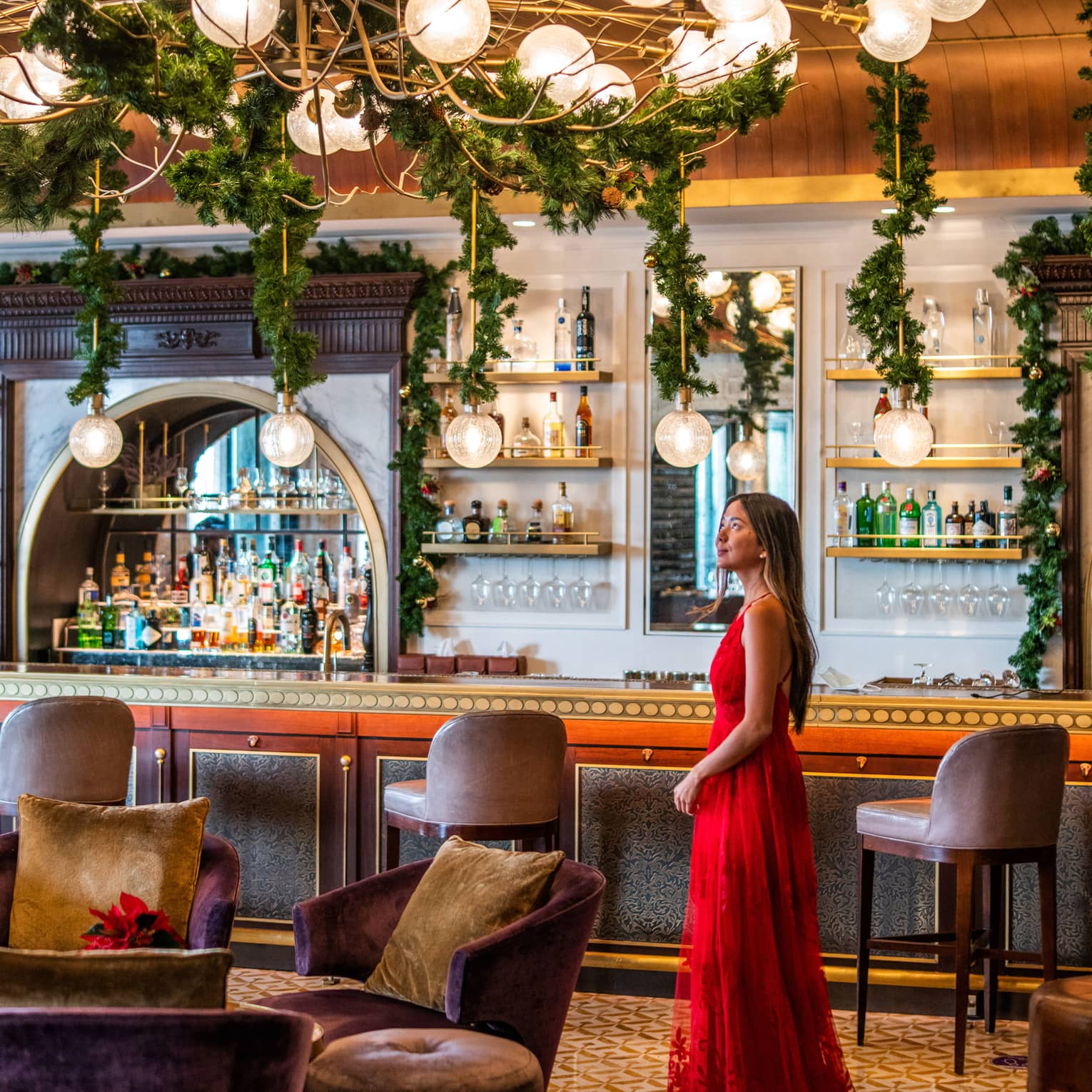 A woman in a long red dress stands in the middle of La Capitale restaurant beneath green festive garlands