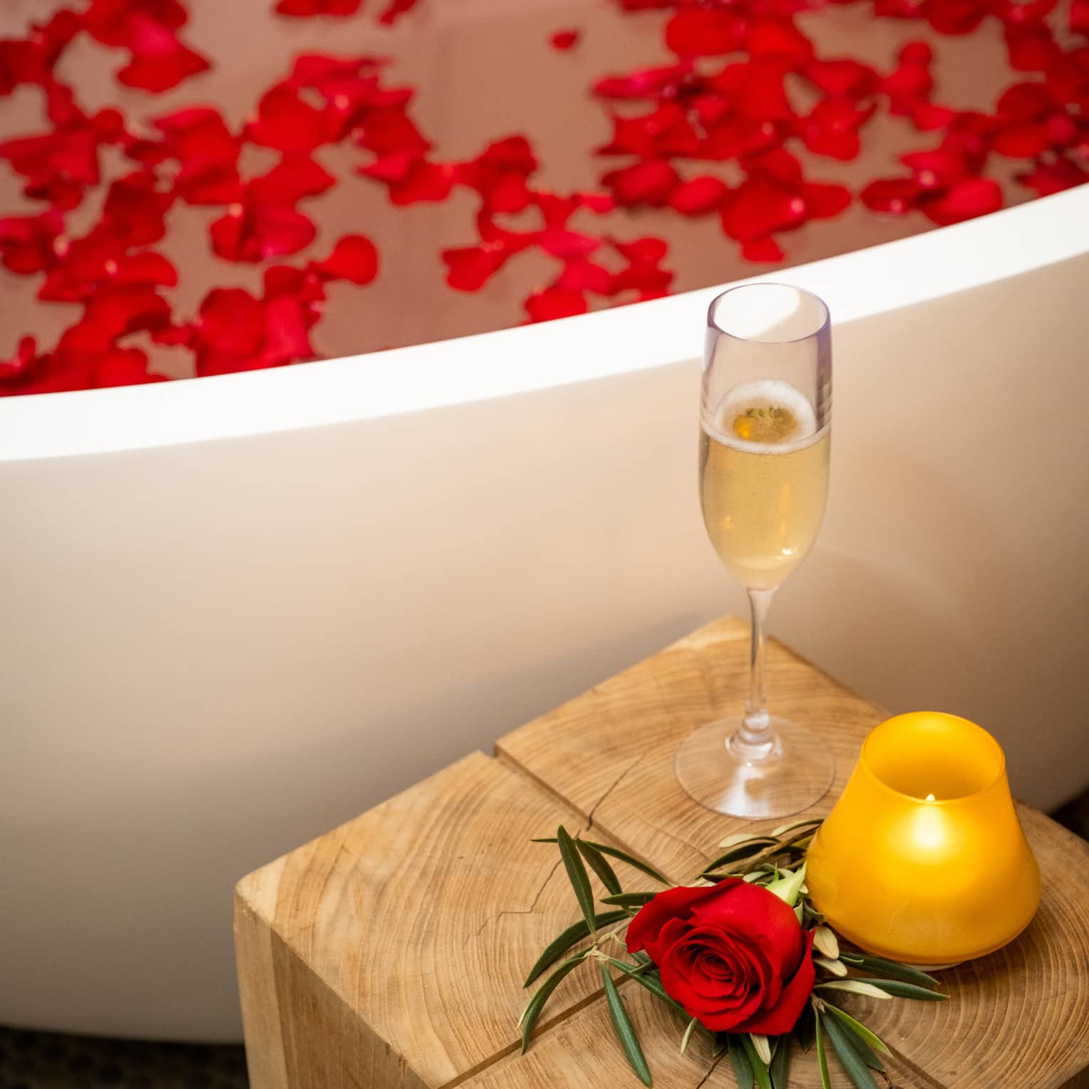 A rose, glass of wine, and candle next to a tub of water and flower petals.