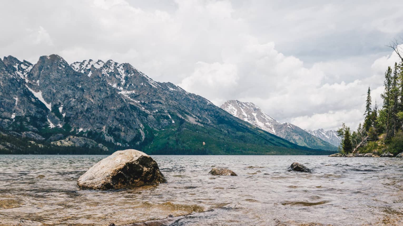 A long view from the pebbled beach looking out into the water with trees lining one shoreline and mountains lining the other.