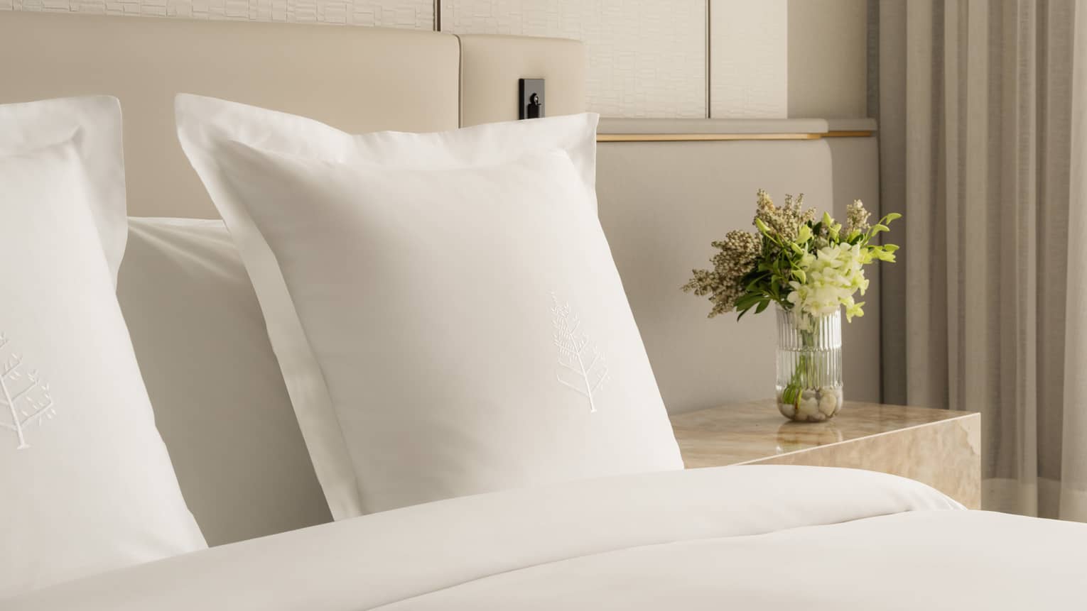 Close-up of a hotel bed with white pillows embroidered with Four Seasons logos and a nightstand with a glass vase of fresh flowers