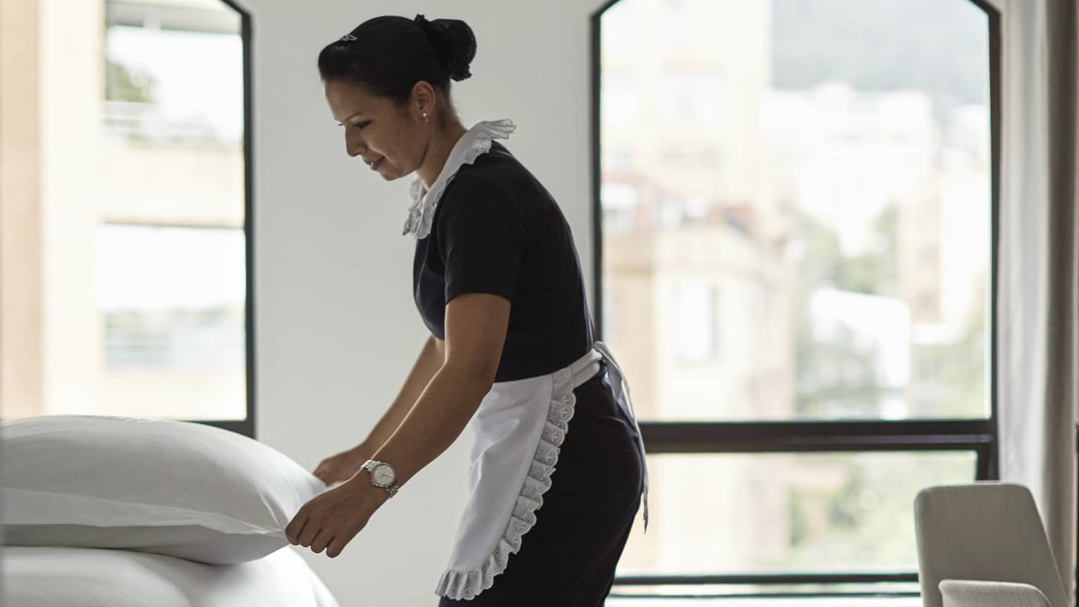 Housekeeping staff makes bed in hotel room by sunny windows 